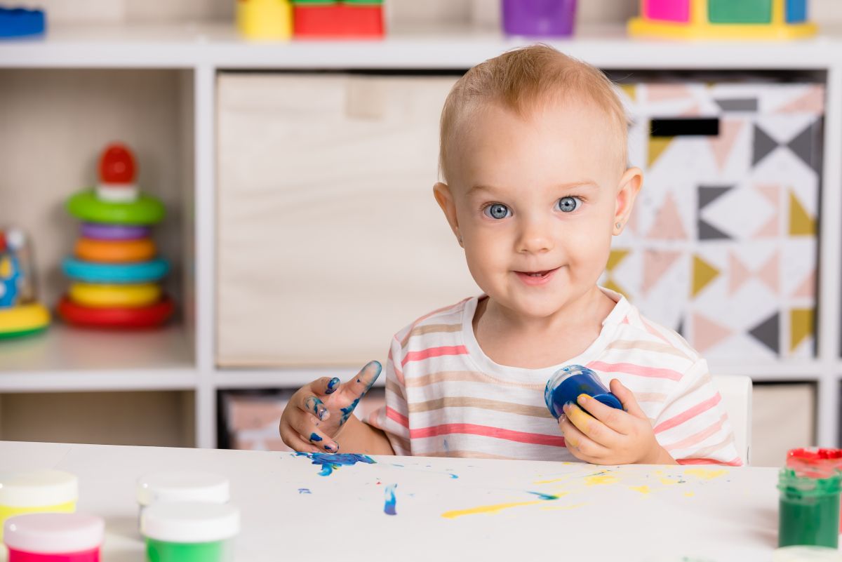 Toddler fetiță cu un tricou cu dungi colorate stă la o masă în dormitor și are în spate un dulab cu etajere pe care stau jucării, ține în mână un borcan cu vopsea albastră, pentru pictură, și este murdar de acea vopsea pe mâna cealaltă, iar masa este pictată cu vopsea albastră și galbenă și mai sunt niște borcane cu vopsea verde, roz, portocalie, ilustrând activități pe care poți să le faci cu toddlerul