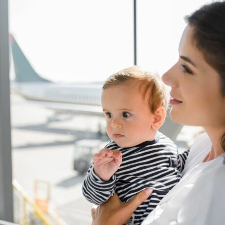 Mamă îmbrăcată cu o bluză albă, care ține în brațe un bebeluș îmbrăcat cu o bluză albă cu dungi negre, stau la poartea de îmbarcare în avion și pe geam se vede coada unui avion