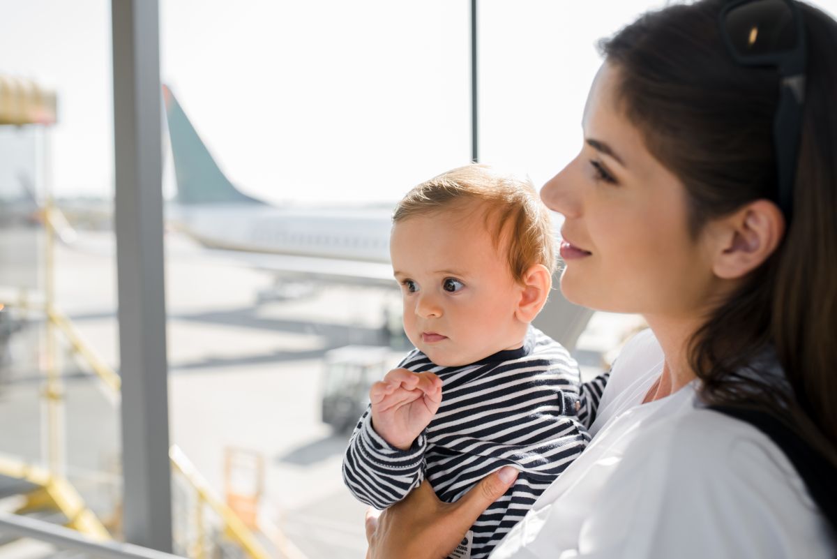 Mamă îmbrăcată cu o bluză albă, care ține în brațe un bebeluș îmbrăcat cu o bluză albă cu dungi negre, stau la poartea de îmbarcare în avion și pe geam se vede coada unui avion