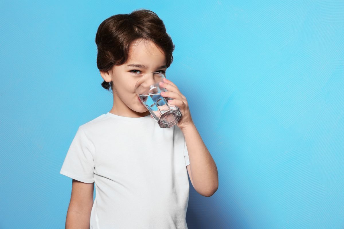 Băiat brunet, îmbrăcat cu tricou alb, care ține în mână un pahar cu apă din care bea, iar în spate are un fundal albastru, ilustrând băuturi sănătoase pentru copii