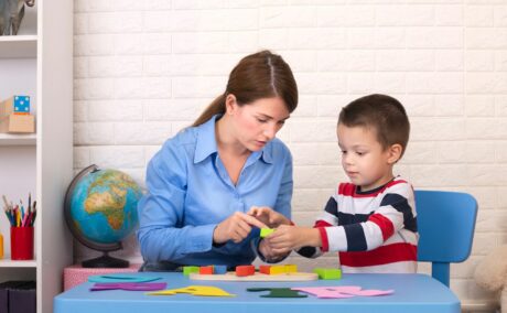 Toddler băiețel, cu o bluză cu dungi groase albe, roșii și bleumarin, stă la o masă din plastic, albastră, alături de logoped, care are o cămașă albastră și învață prin jocul cu cuburi colorate, dar și litere mari, iar în spate se vede un dulap cu diferite obiecte și un glob pământesc