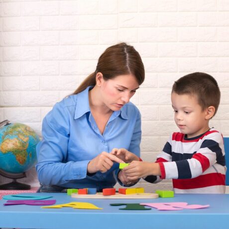 Toddler băiețel, cu o bluză cu dungi groase albe, roșii și bleumarin, stă la o masă din plastic, albastră, alături de logoped, care are o cămașă albastră și învață prin jocul cu cuburi colorate, dar și litere mari, iar în spate se vede un dulap cu diferite obiecte și un glob pământesc