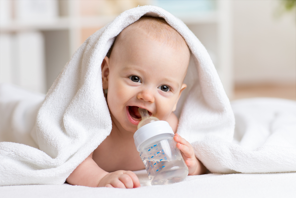 Un bebeluș care bea apă dintr-un biberon, acoperit de un prosop de baie