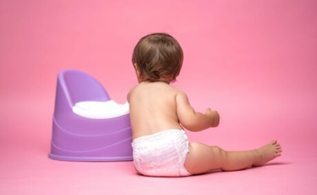 Bebeluș cu scutec fără haine, care stă așezat în fund, pe podea, cu spatele, sprijinit cu un cot de o oliță mov