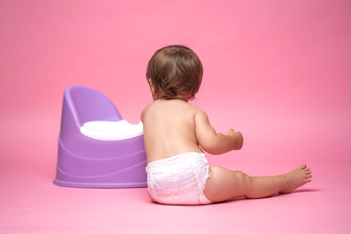 Bebeluș cu scutec fără haine, care stă așezat în fund, pe podea, cu spatele, sprijinit cu un cot de o oliță mov