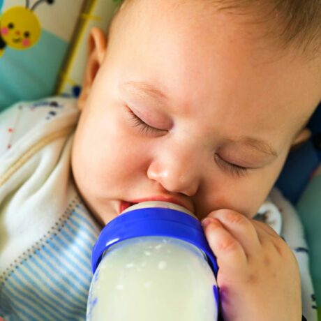 Bebeluș care doarme într-un scaun cu albinuțe și ține în gură un biberon din sticlă, cu lapte, cu o parte din plastic albastru