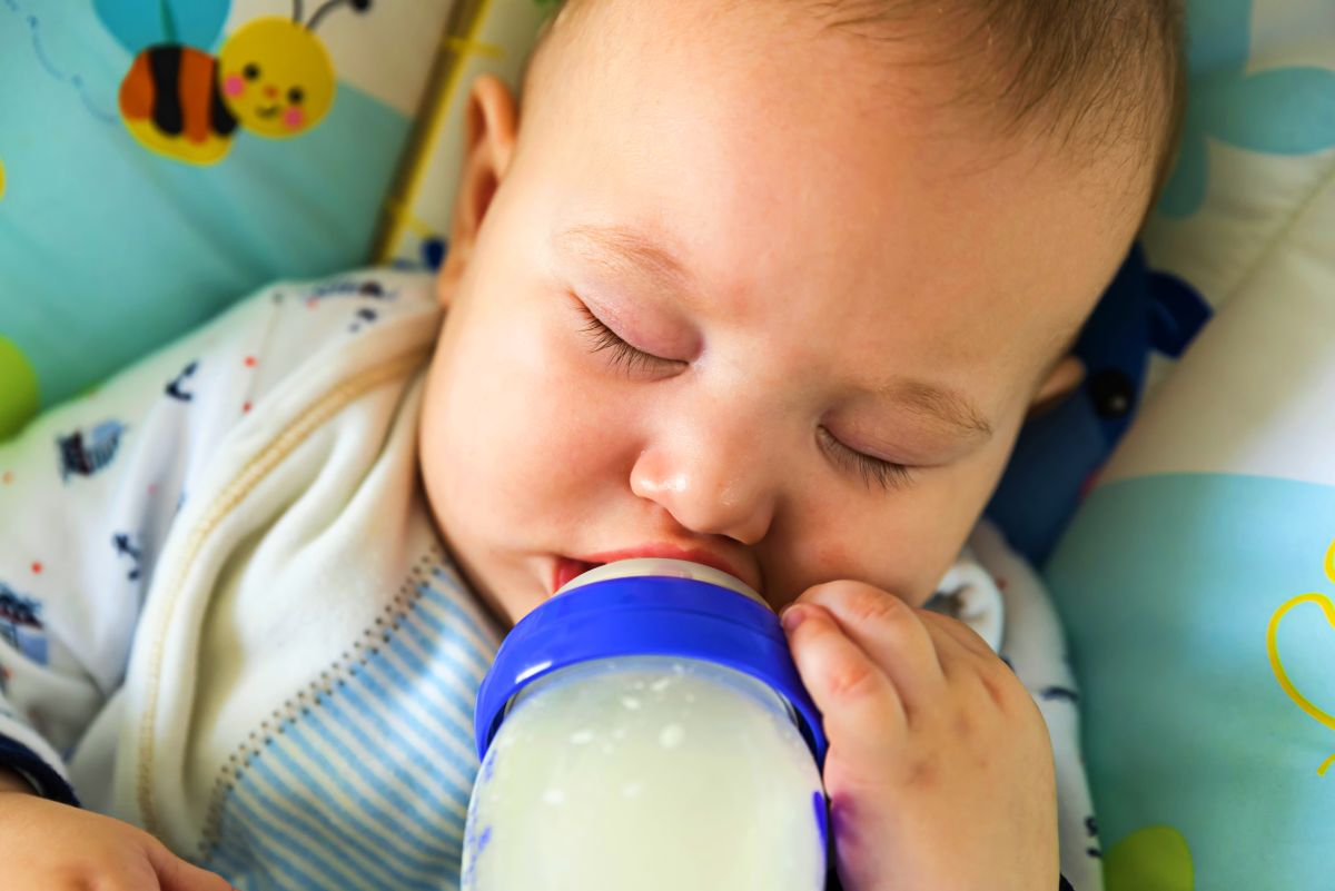 Bebeluș care doarme într-un scaun cu albinuțe și ține în gură un biberon din sticlă, cu lapte, cu o parte din plastic albastru