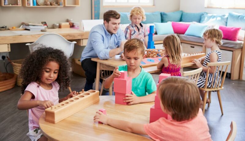 Profesor într-o sală de clasă Montessori și copii așezați la mese din lemn, jucându-se cu cuburi și jucării din lemn