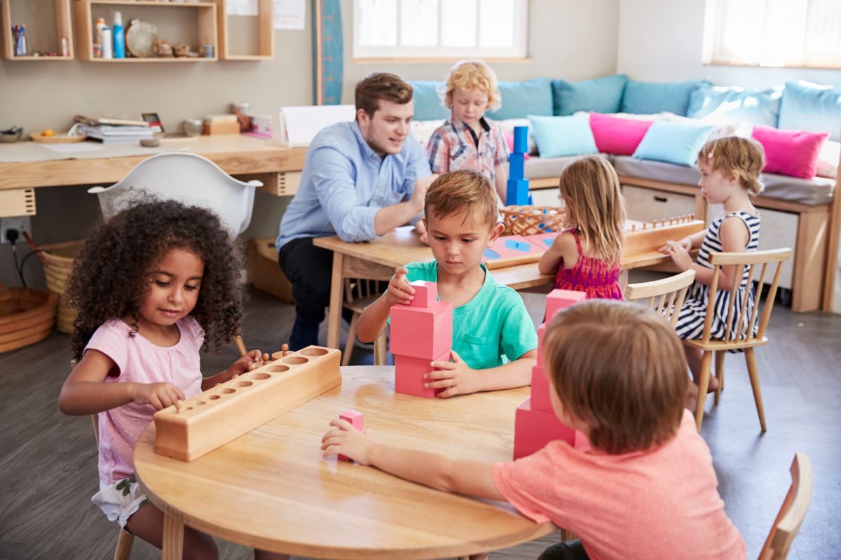Profesor într-o sală de clasă Montessori și copii așezați la mese din lemn, jucându-se cu cuburi și jucării din lemn