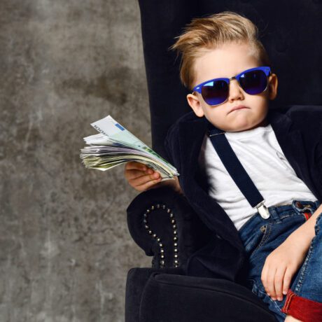 Un băiețel aranjat, sacou, ochelari de soare și mulți bani într-o mână, pe un fotoliu masiv