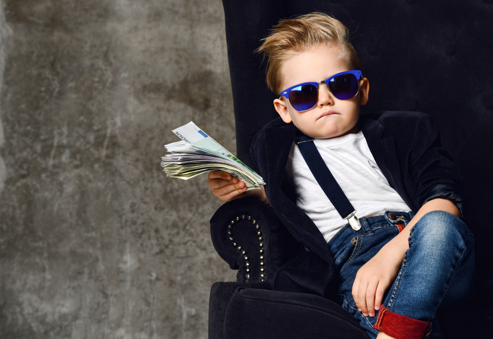 Un băiețel aranjat, sacou, ochelari de soare și mulți bani într-o mână, pe un fotoliu masiv