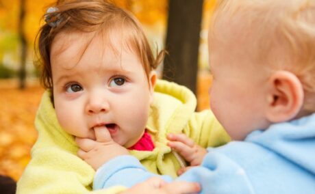 O fețită bebeluș, cu un hanorac galbe, mușcă degetul unui băiețel cu hanorac bleu, afară în parc, toamna