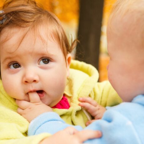 O fețită bebeluș, cu un hanorac galbe, mușcă degetul unui băiețel cu hanorac bleu, afară în parc, toamna