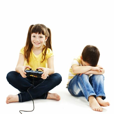 Doi copii, o fetiță cu tricou galben și jeanși care are în mână o telecomandă pentru joc și e fericită, iar băiețelul de lângă ea este supărat și stă cu genunchii ridicați și capul sprijinit peste aceștia, ilustrând copilul tău nu știe să piardă