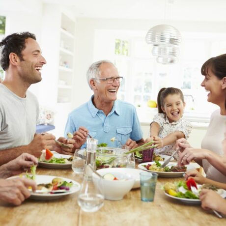 Familie, formată din părinți, bunici și copii, stau la masă, mănâncă și râd, iar pe lângă farfurii și boluri cu mâncare sunt și pahare cu apă