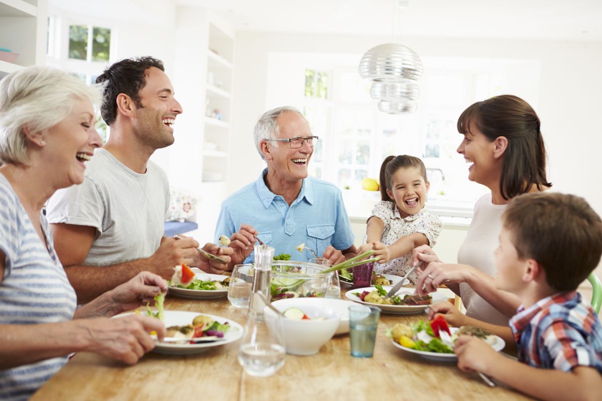 Familie, formată din părinți, bunici și copii, stau la masă, mănâncă și râd, iar pe lângă farfurii și boluri cu mâncare sunt și pahare cu apă