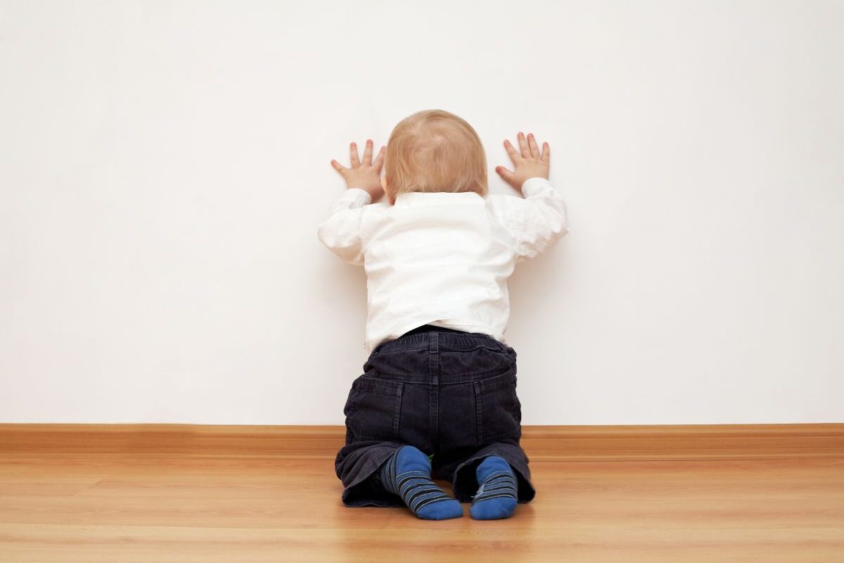 Bebeluș care stă cu spatele, în genunchi, cu mâinile sprijinite de perete și își lovește capul, îmbrăcat cu o cămașă albă, pantaloni bleumarin și șosete albastre cu dungi bleumarin