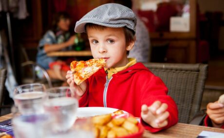 Băiețel, îmbrăcat cu tricou galben, hanorac roșu pe deasupra și o șapcă cu dungi albe și negre, ține într-o mână, pe care o duce la gură, o felie de pizza, stând pe un scaun la restaurant, iar pe masă mai sunt niște pahare cu apă și un bol cu cartofi prăjiți