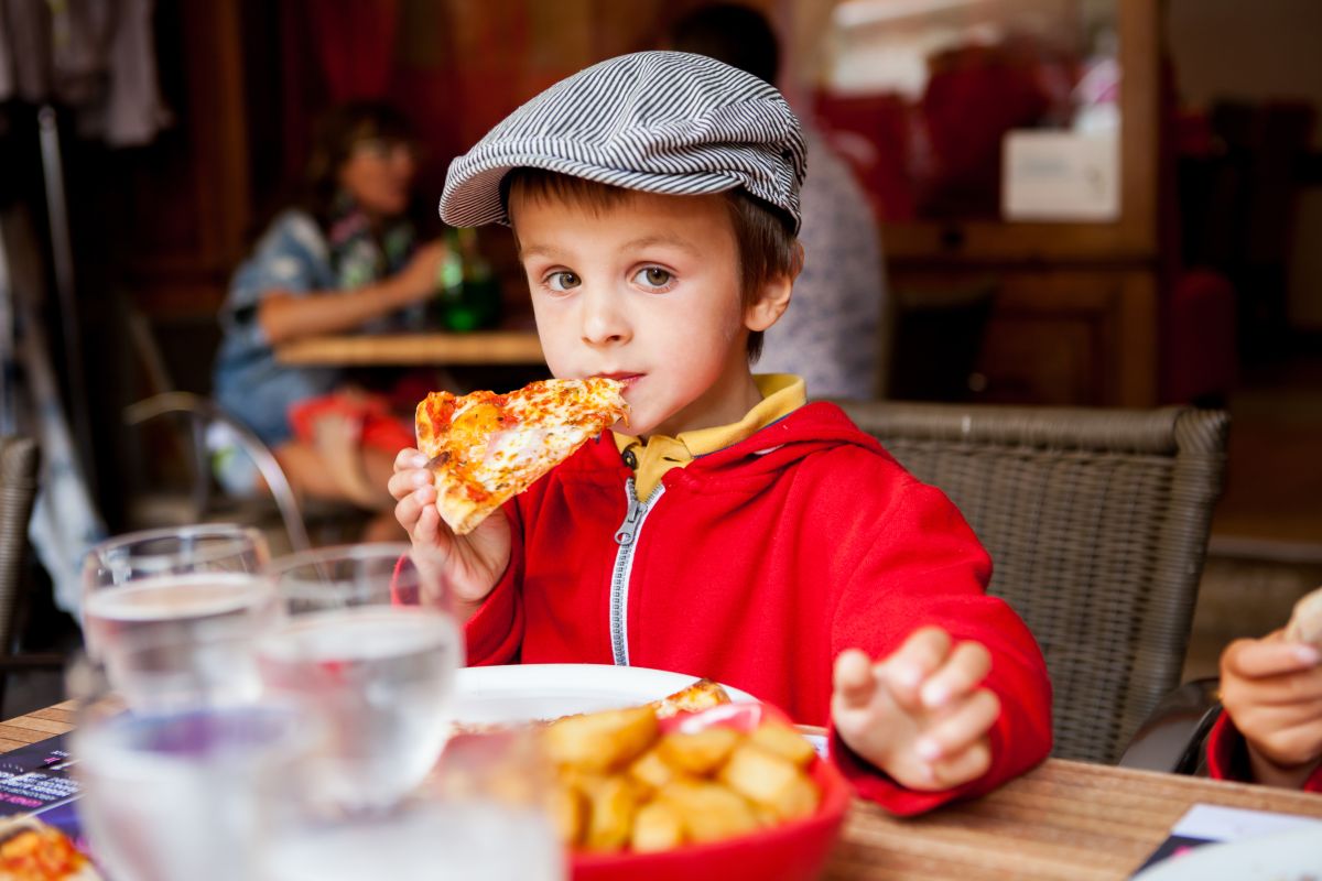 Băiețel, îmbrăcat cu tricou galben, hanorac roșu pe deasupra și o șapcă cu dungi albe și negre, ține într-o mână, pe care o duce la gură, o felie de pizza, stând pe un scaun la restaurant, iar pe masă mai sunt niște pahare cu apă și un bol cu cartofi prăjiți