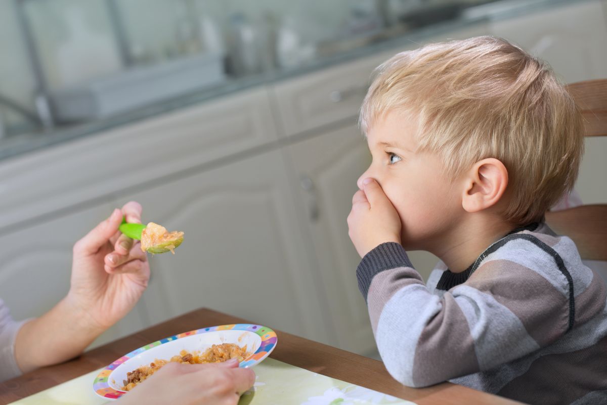 Băiețel blond, stă la masă și își ține mâna la gură în semn că refuză lingura verde, cu carne, pe care i-o oferă mama lui, care ține în cealaltă mână farfuria cu tocăniță cu carne, ilustrând copilul refuză carnea