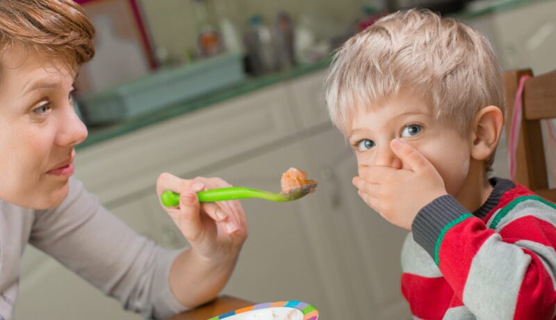 Mama care stă cu copilul la masă, în bucătărie, și îi oferă copilului mâncare într-o linguriță verde, iar acesta o refuză, punând mâna la gură, ilustrând de ce sunt copiii mofturoși la masă