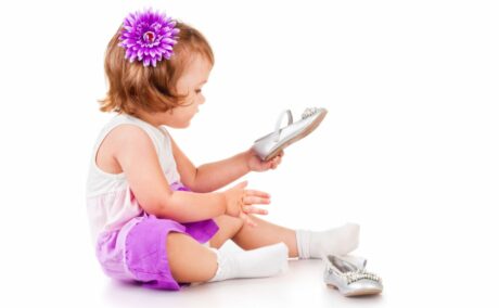 Fetiță îmbrăcată cu o rochiță alb cu mov și șosete albe, cu o floare mov prinsă în păr, încearcă să își puna un pantof gri în picioare, iar celălalt pantof este lângă ea