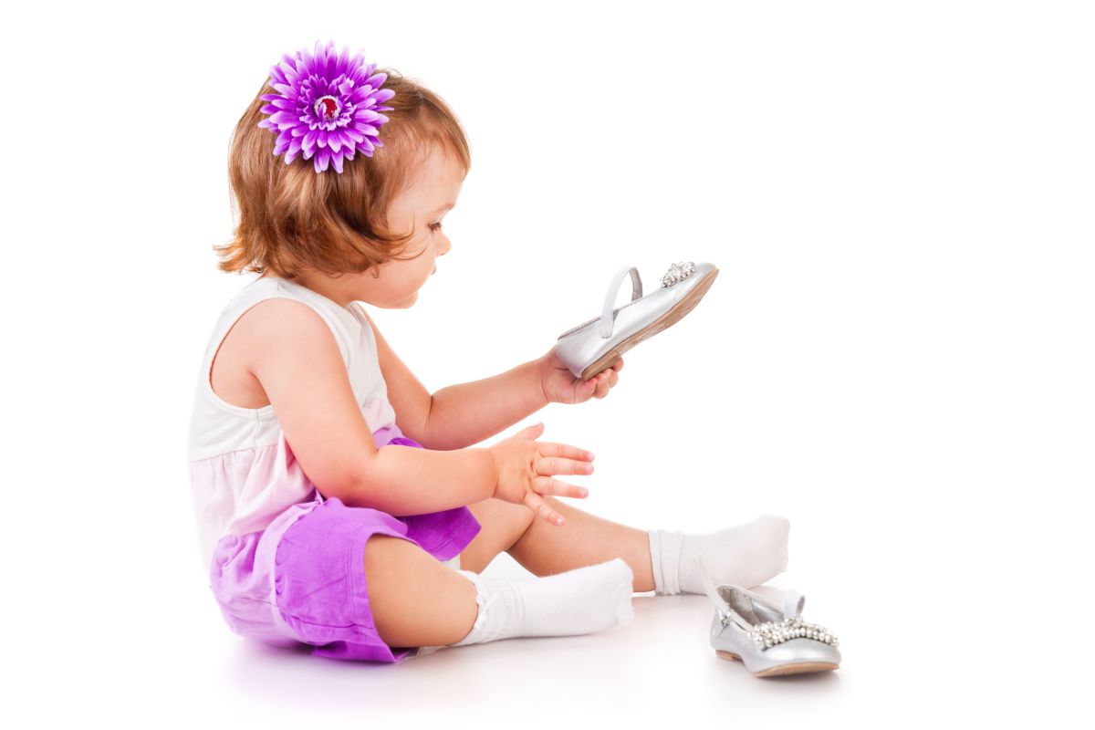 Fetiță îmbrăcată cu o rochiță alb cu mov și șosete albe, cu o floare mov prinsă în păr, încearcă să își puna un pantof gri în picioare, iar celălalt pantof este lângă ea