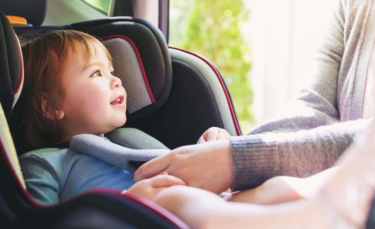 Toddler care stă în scaunul de mașină, poziționat cu fața la sensul de mers, iar mama lui îi prinde centurile