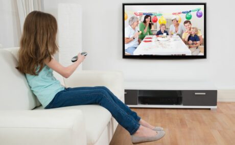 Fetiță îmbrăcată cu un tricou bleu, jeanși și balerini, care stă pe o canape, în living, cu o telecomandă în mână și se uită la televizor, la o petrecere aniversară a unui copil, ilustrând cum să reduci timpul de privit la ecrane