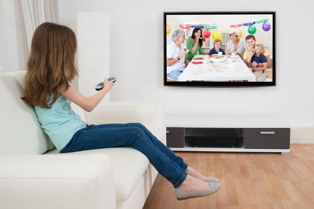 Fetiță îmbrăcată cu un tricou bleu, jeanși și balerini, care stă pe o canape, în living, cu o telecomandă în mână și se uită la televizor, la o petrecere aniversară a unui copil, ilustrând cum să reduci timpul de privit la ecrane