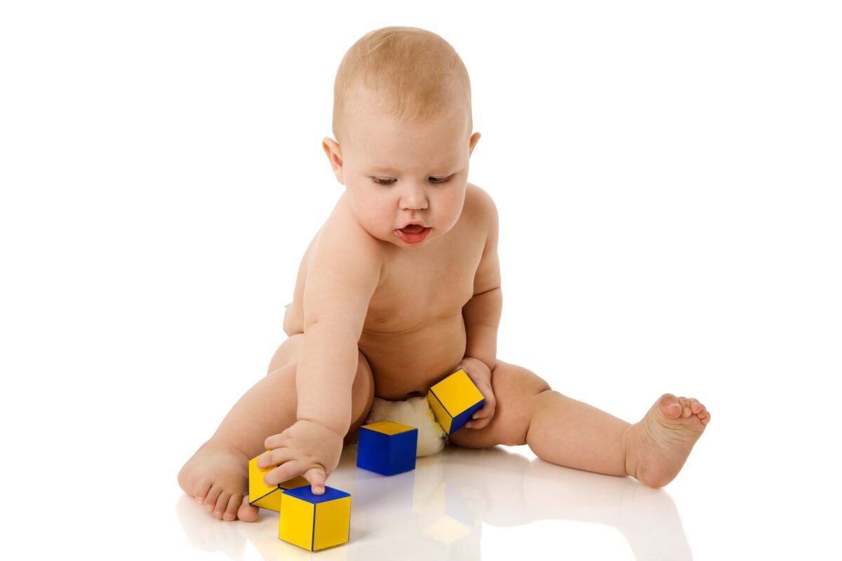 Bebeluș care stă în fund, dezbrăcat, doar în scutec, și se joacă cu cuburi colorate, galben cu abastru, ilustrând dezvoltarea motricității fine la bebeluși