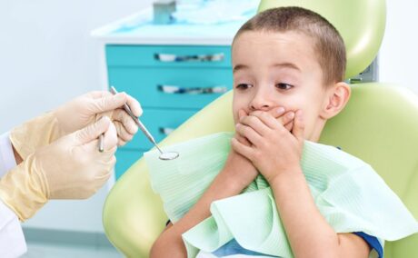 Băiețel pe scaunul din cabinetul stomatologic, care are o bavetă verde la gât și își ascunde gura cu mâinile