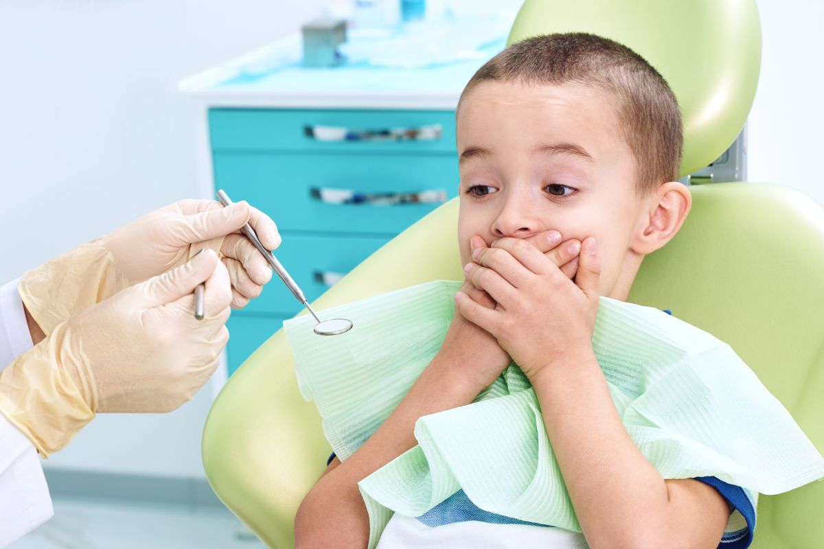 Băiețel pe scaunul din cabinetul stomatologic, care are o bavetă verde la gât și își ascunde gura cu mâinile