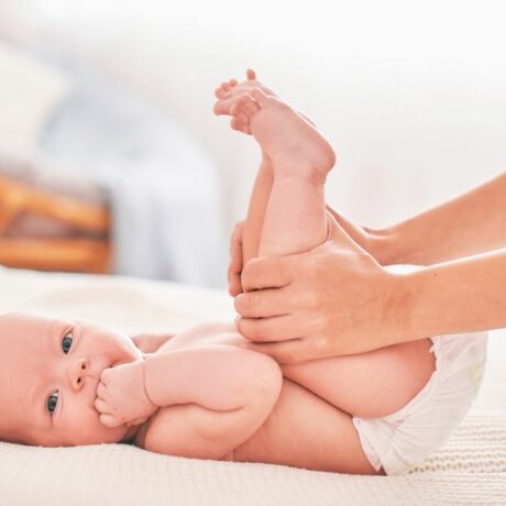 Bebeluș care stă pe spate, pe un pat cu cearșaf alb, în dormitor, doar cu scutec alb pe el, iar mama lui îi ține picioarele ridicate și îl ajută să pedaleze, ilustrând gazele intestinale la bebeluși