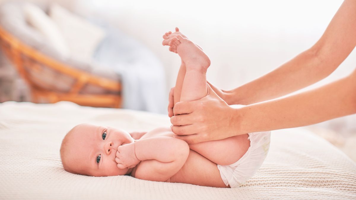 Bebeluș care stă pe spate, pe un pat cu cearșaf alb, în dormitor, doar cu scutec alb pe el, iar mama lui îi ține picioarele ridicate și îl ajută să pedaleze, ilustrând gazele intestinale la bebeluși