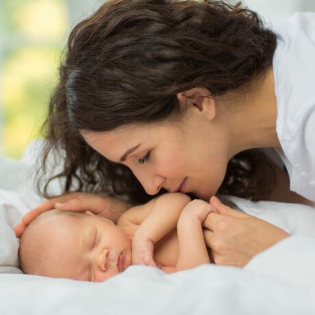 Mamă brunetă cu părul lung și ondulat, aplecată asupra bebelușului care stă pe un pat cu lenjerie albă, cu o mână îi mângâie capul și cu cealaltă mână îi ține o mânuță și îi sărută brațul, ilustrând greșeli în îngrijirea bebelușului