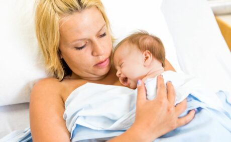 Proaspătă mamă, care stă pe patul de spital,acoperită cu un cearșaf, și ține la piept bebelușul care plânge.