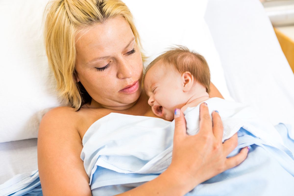Proaspătă mamă, care stă pe patul de spital,acoperită cu un cearșaf, și ține la piept bebelușul care plânge.