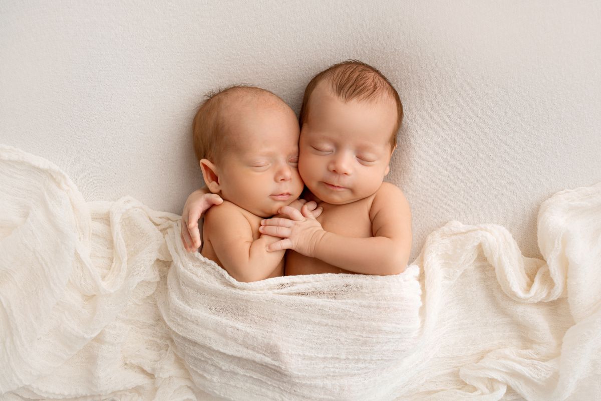 Bebeluși, gemeni, care se stau îmbrățișați și unt acoperiți cu o eșarfă albă