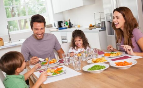 Familie veselă, cu 2 copii, la masă, în bucătăărie, mâncând mâncare sănătoasă și având în pahare apă
