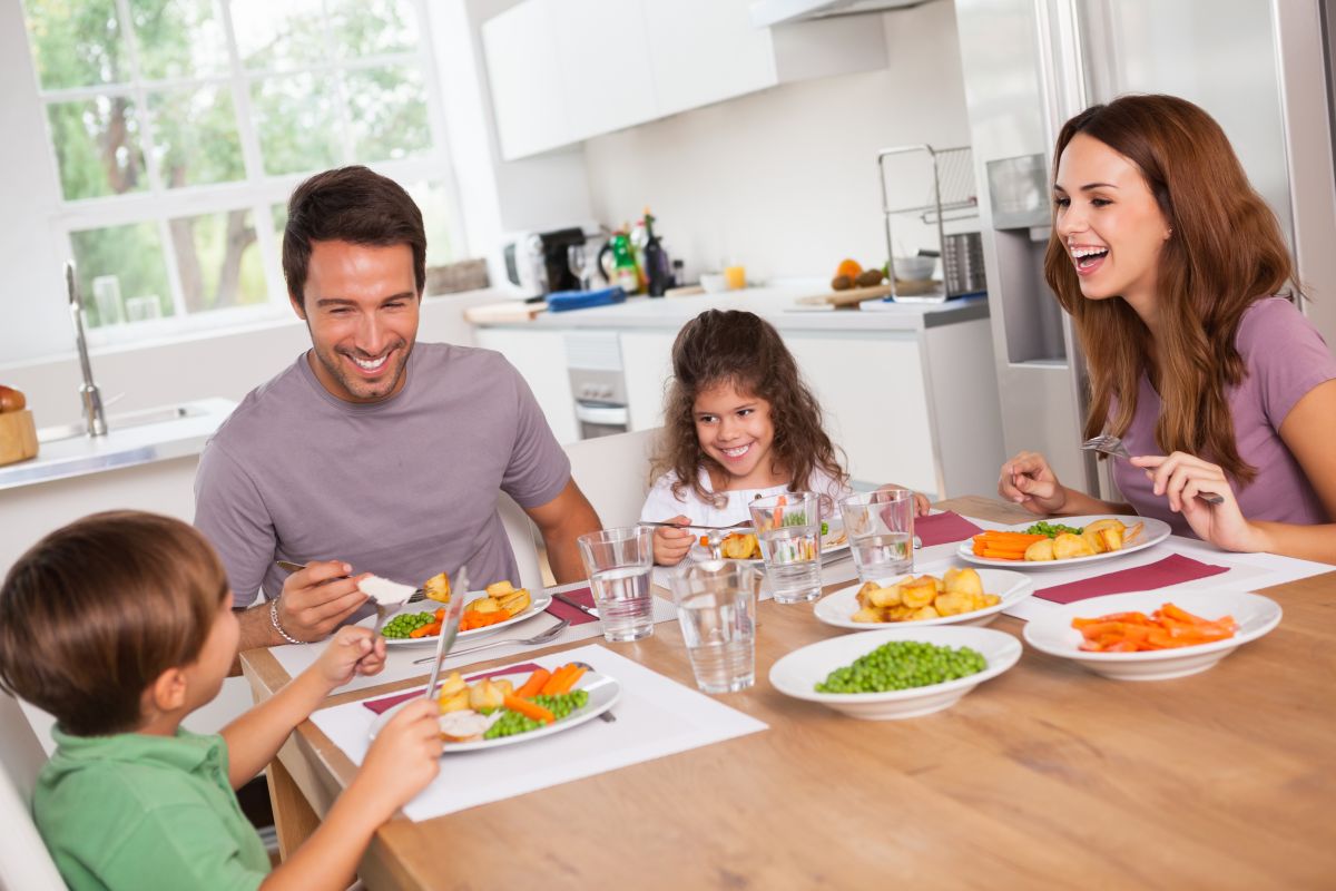 Familie veselă, cu 2 copii, la masă, în bucătăărie, mâncând mâncare sănătoasă și având în pahare apă