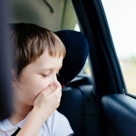 Băiețel, îmbrăcat cu un tricou alb, care stă în scunul de mașină și se ține cu mâna de gură și nas pentru că are rău de mașină