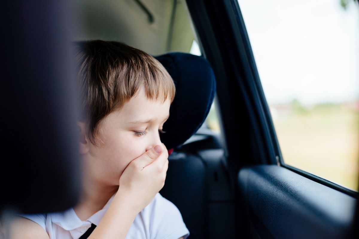 Băiețel, îmbrăcat cu un tricou alb, care stă în scunul de mașină și se ține cu mâna de gură și nas pentru că are rău de mașină