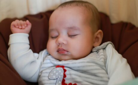 Bebeluș care doarme pe spate, pe o pătură maro, îmbrăcat cu un body gri cu dinozaur, și stă cu o mână ridicată și are urme de reflux gastroesofagian la gură