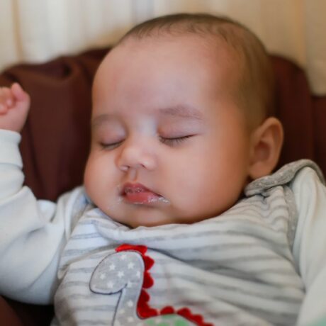 Bebeluș care doarme pe spate, pe o pătură maro, îmbrăcat cu un body gri cu dinozaur, și stă cu o mână ridicată și are urme de reflux gastroesofagian la gură