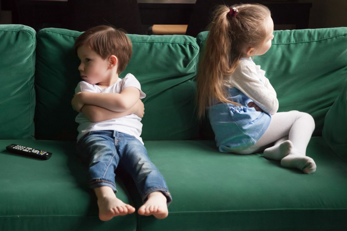 Doi frați care stau supărați pe o canapea verde, băiețelul stă cu fața și cu mâinile încrucișate și fetița stă întoarsă cu spatele
