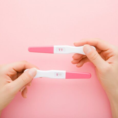 Două mâini în care sunt ținute 2 teste de sarcină, roz cu alb, unul negativ și unul pozitiv, pe un fundal roz pal