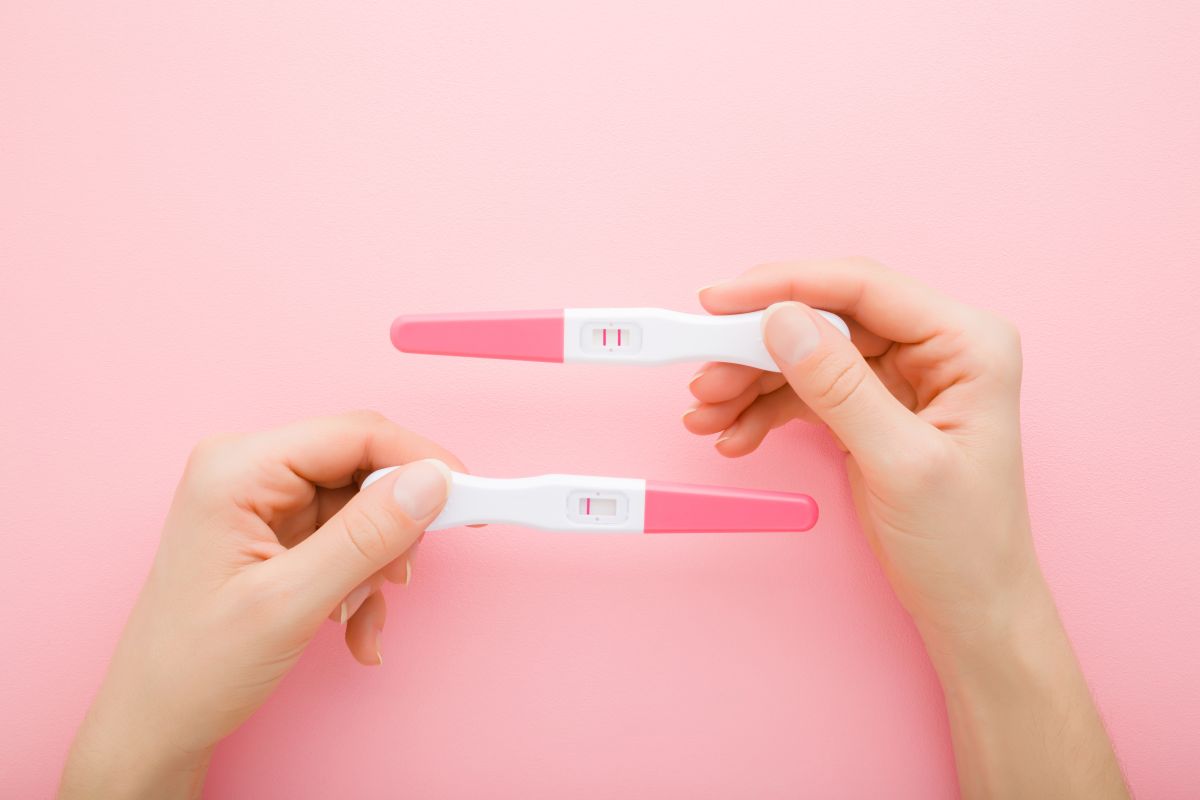 Două mâini în care sunt ținute 2 teste de sarcină, roz cu alb, unul negativ și unul pozitiv, pe un fundal roz pal