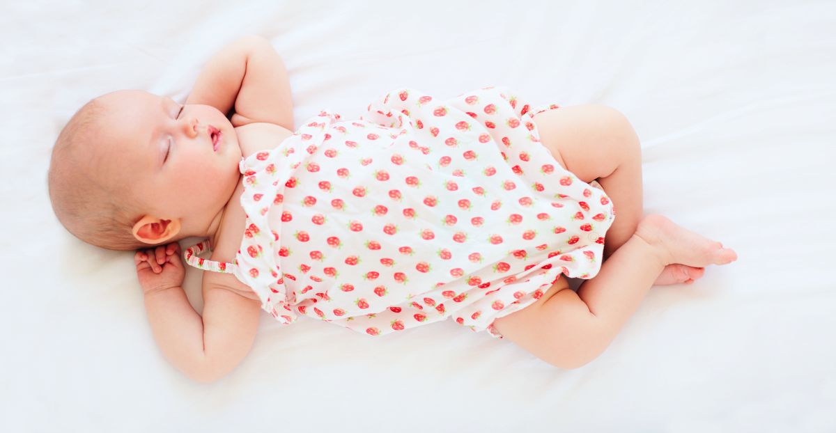 Bebeluș fetiță, care doarme pe spate, cu mâinile sub cap și picioarele încrucișate, pe un cearșaf alb și este îmbrăcată cu un body cu bretele, alb cu căpșuni