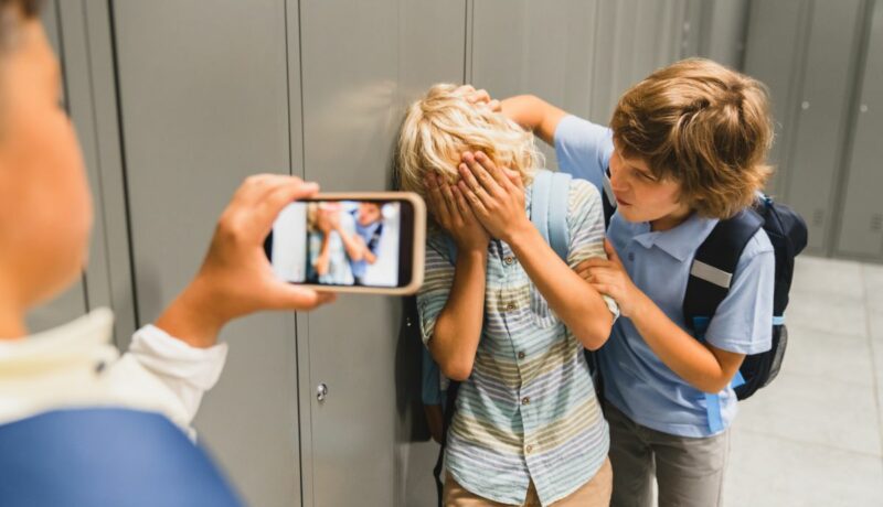 Bullying cibernetic, în care un copil filmează în timp ce un copil îl agresează pe altul prinzândul din spate de o mână și de gât, iar celălat se apără ținând mainile la față